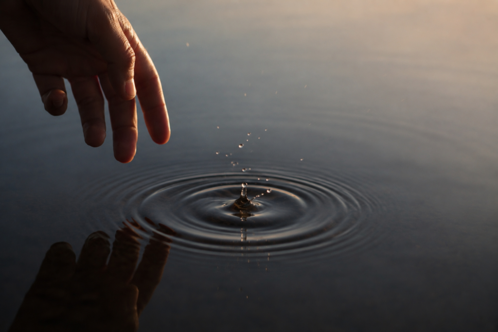 Un doigt effleure une eau calme et fait naître des ondes concentriques qui se propagent au loin.