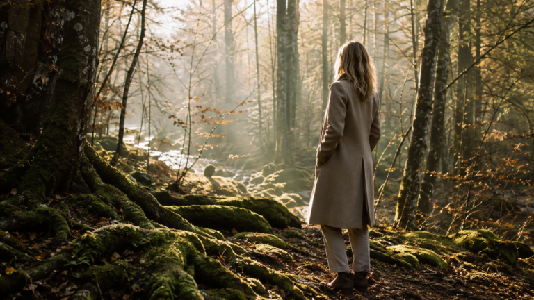 Une femme élégante en manteau, de dos, contemple une forêt lumineuse au petit matin, où racines moussues et ruisseau tissent le sol : image de la conscience des liens en Sageocratie.